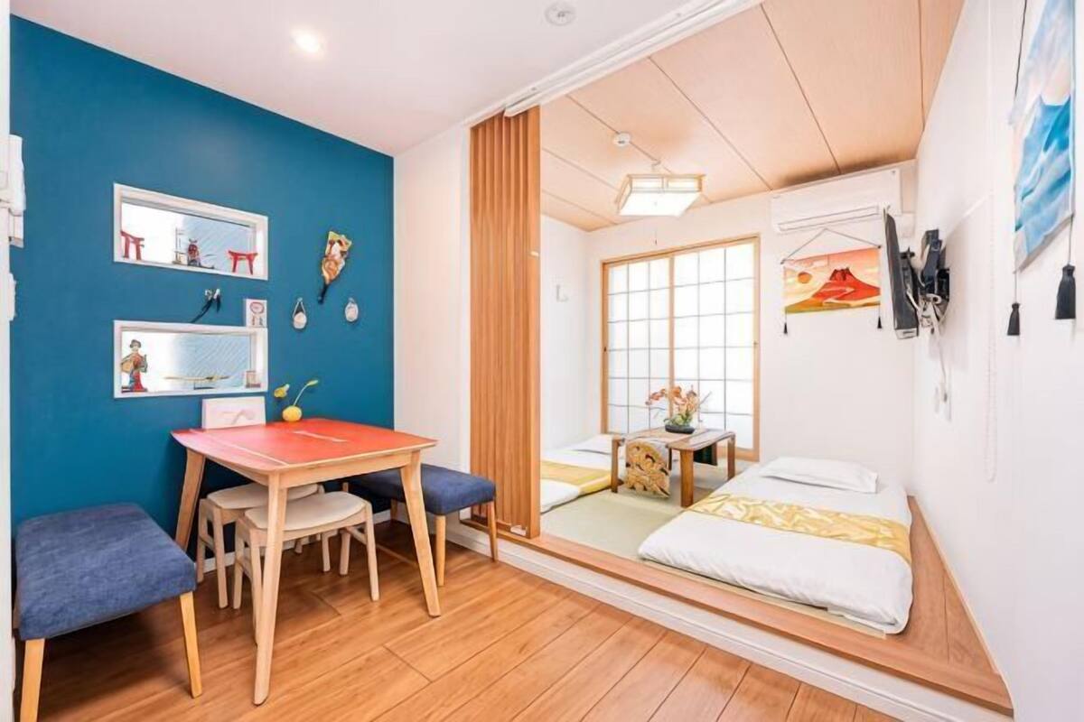 Une pièce avec un plancher en bois, un lit recouvert d’une courtepointe blanche, une petite table avec des chaises et un mur bleu orné de décorations.