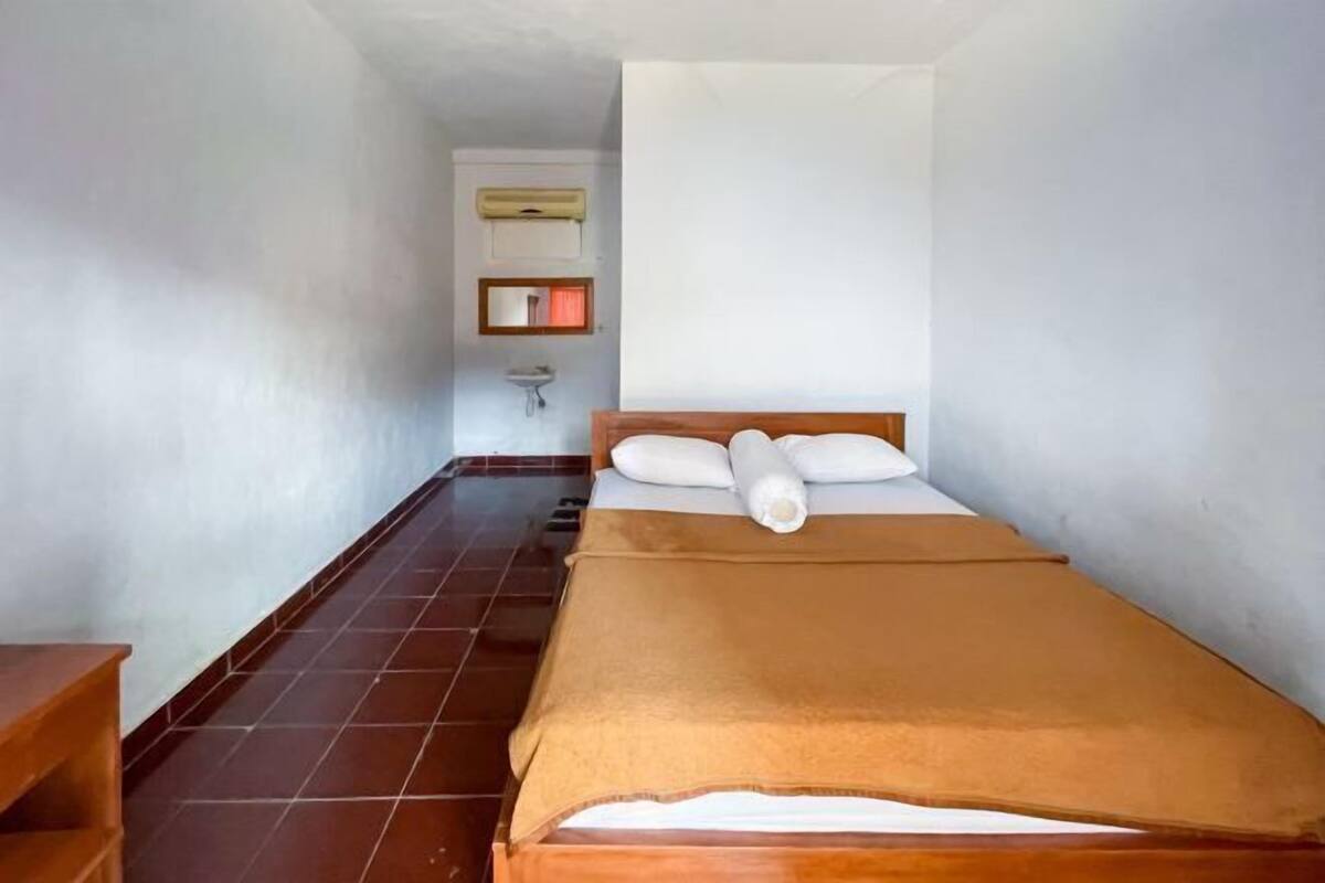 A small, narrow room with a single bed, a wooden nightstand, a wall-mounted sink, and a tiled floor.
