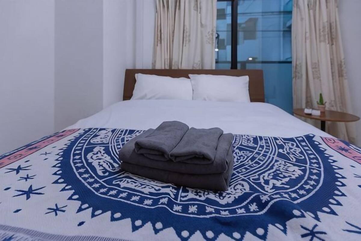 A neatly made bed with a patterned blanket, a bedside table with a plant, and a window with curtains.