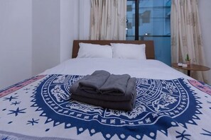 Een opgemaakt bed met een geborduurd plaid, een nachtkastje met een plant en een raam met gordijnen.