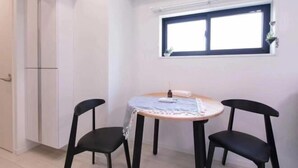A small, round dining table with two black chairs, a window with blinds, and a white wall. - japan house sugamo (Tokyo)