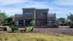 A modern two-story building with a flat roof, large windows, and a covered outdoor seating area with green chairs. - Thechaeum (Seogwipo)