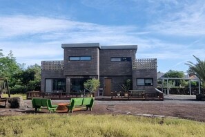 A modern two-story building with a flat roof, large windows, and a covered outdoor seating area with green chairs. - Thechaeum (Seogwipo)