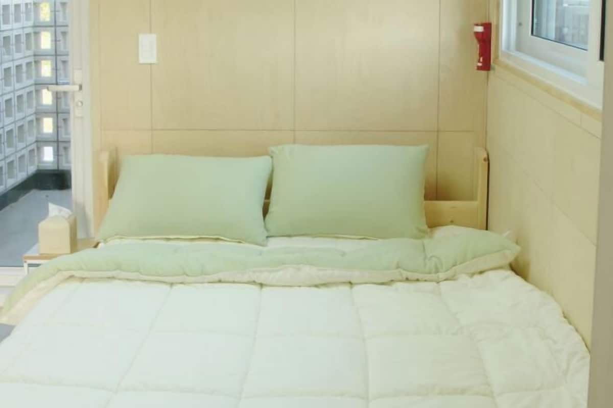 A bed with a quilted mattress and two pillows, a window with a view of a building, and a wall-mounted light switch.