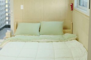 A bed with a quilted mattress and two pillows, a window with a view of a building, and a wall-mounted light switch. - Thechaeum (Seogwipo)