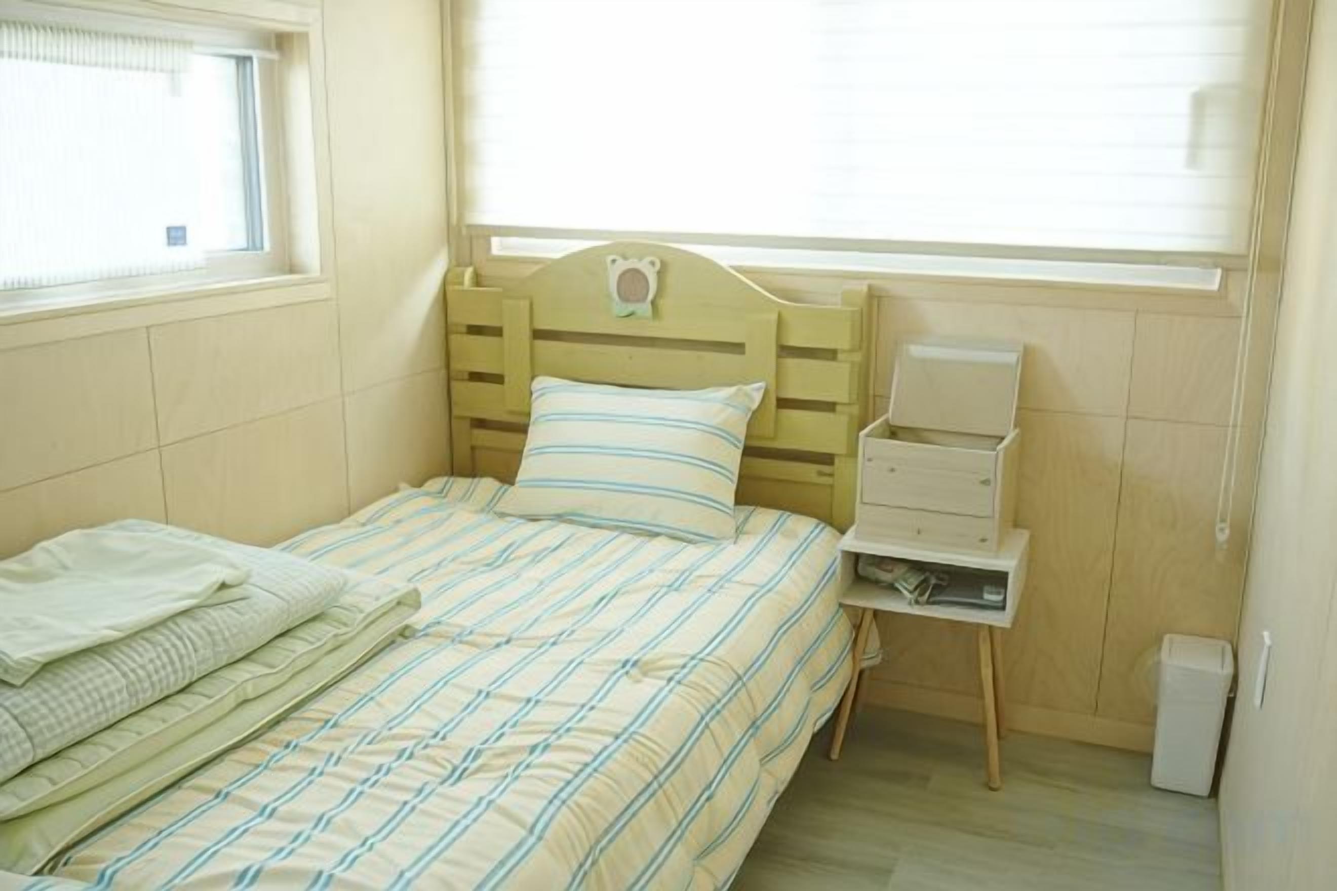 A single bed with a headboard, a bedside table, and a window with blinds.
