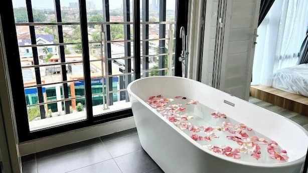 A bathroom with a bathtub, a view through large windows, and a bed with a white bedspread.
