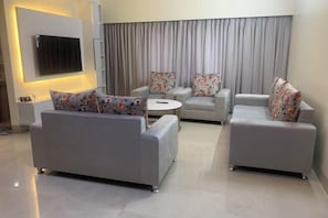A modern living room with a grey sofa, two armchairs, a small round table, and a flat-screen TV mounted on the wall. - Mahir's Cozy Den (Dhaka)