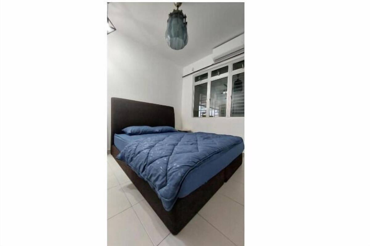 Een bed met een donker hoofdeinde en een blauwe quilt, een raam met zonwering, en een hanglamp.