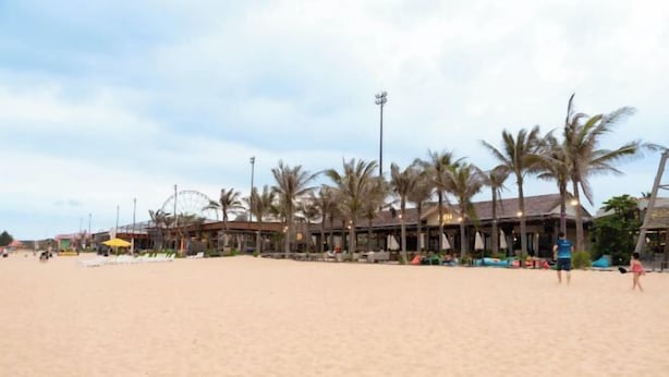 A beach with palm trees, a building with a sloped roof, and a few people walking. - Chang Luxury Villa Beach View (Phan Thiet)