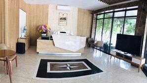A modern reception area with a reception desk, a large flat-screen TV, and a view of greenery through large windows.