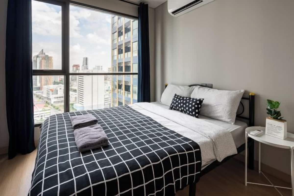 Ein Hotelzimmer mit großem Fenster, einem Bett mit schwarz-weiß kariertem Überwurf und einem kleinen Tisch mit einer Pflanze.