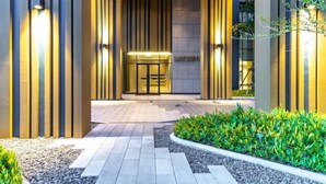 A modern hotel entrance with vertical wooden panels, a glass door entrance, and a landscaped garden area. - Nana Sukhumvit Bangkok Siam Apartment (Bangkok)