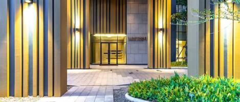 Fachada de hotel con diseño moderno, destacando una puerta de cristal, una zona de recepción y un jardín cuidado.