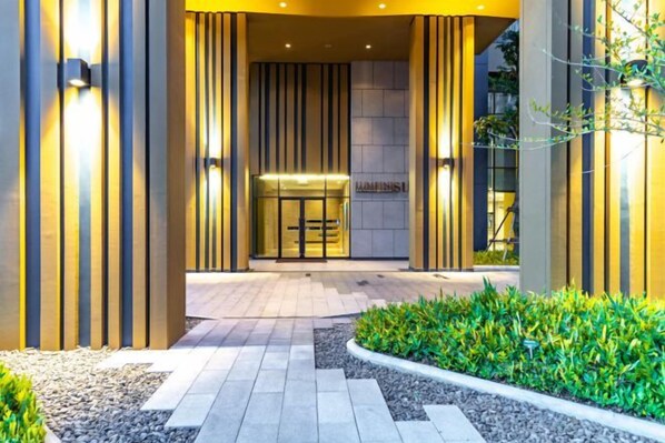 A hotel entrance with a modern design, featuring a glass door, a reception area, and a well-maintained garden.