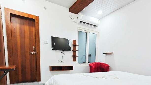 A bedroom with a wooden bed frame, a flat-screen TV mounted on the wall, a window with a view, and a red chair. - Hotel Royal Krishna (Gorakhpur)