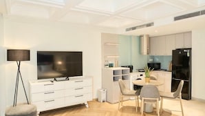 Een moderne woonkamer met een flatscreen televisie, een eethoek met een ronde tafel, en een keuken met witte kasten en een zwarte koelkast.