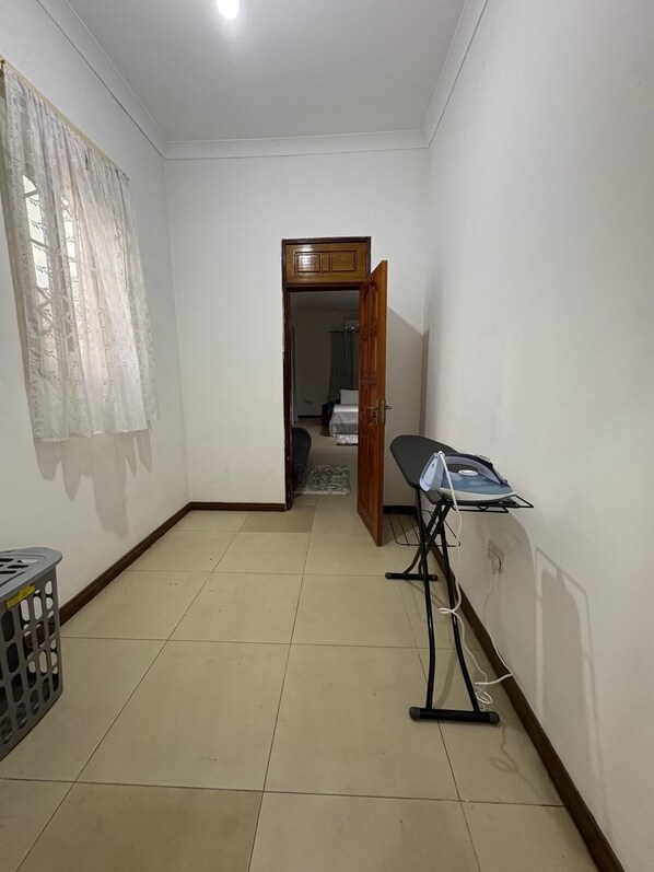 A room with a tiled floor, a wooden door, a laundry basket, and an ironing board.