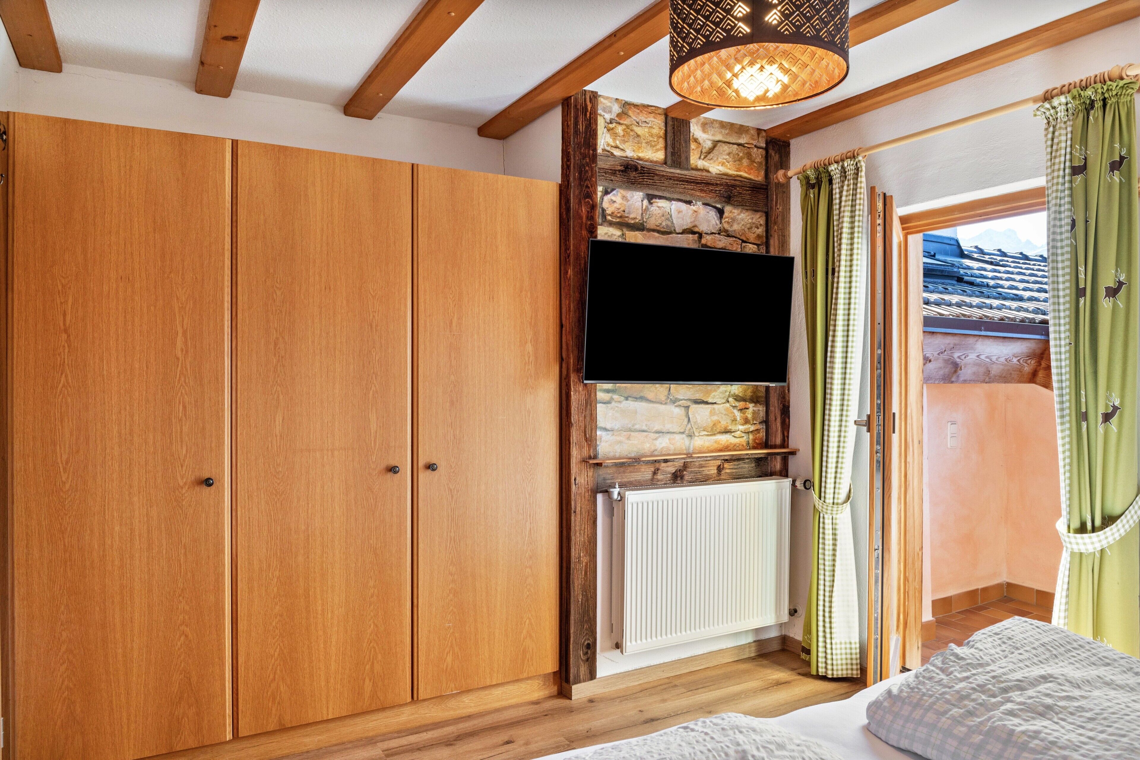 A bedroom with wooden wardrobe, a mounted TV, stone wall, and a view of a mountain through a window.