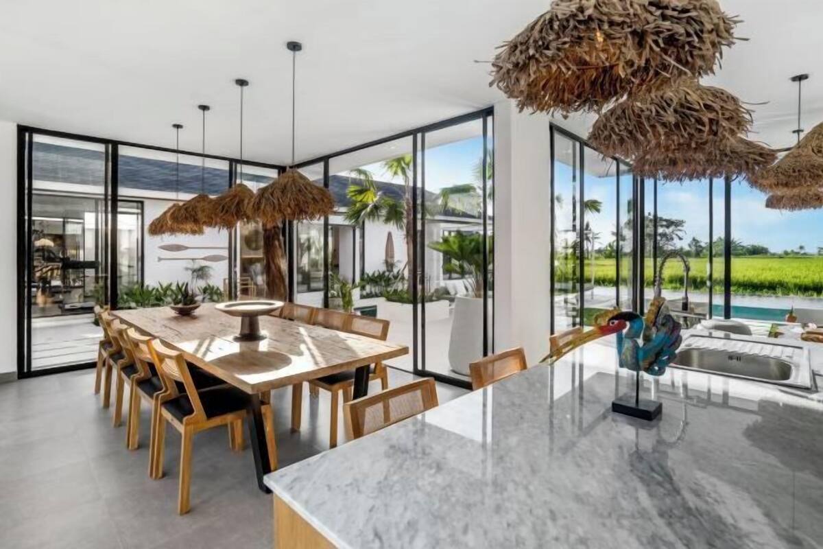 Un espace repas moderne avec un îlot en marbre, des chaises en bois et une vue sur une piscine et un jardin luxuriant.