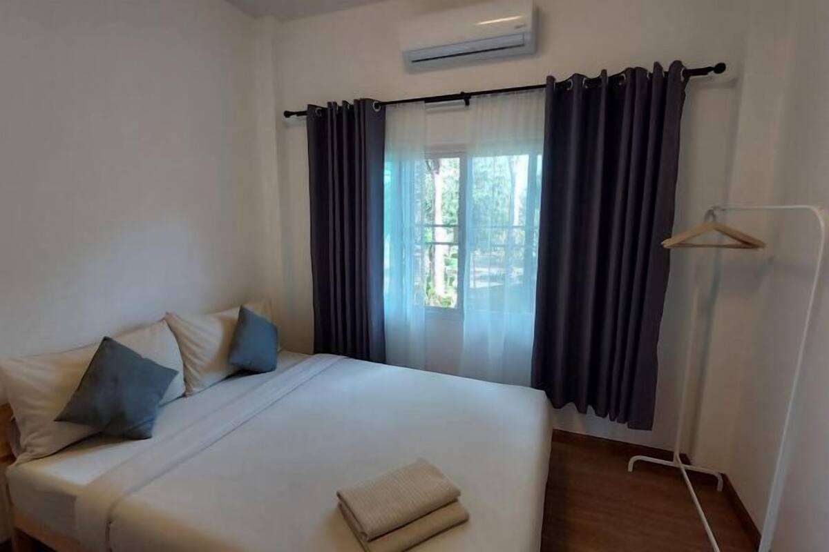 A hotel room with a bed, a window with curtains, a wall-mounted air conditioner, and a clothing rack.