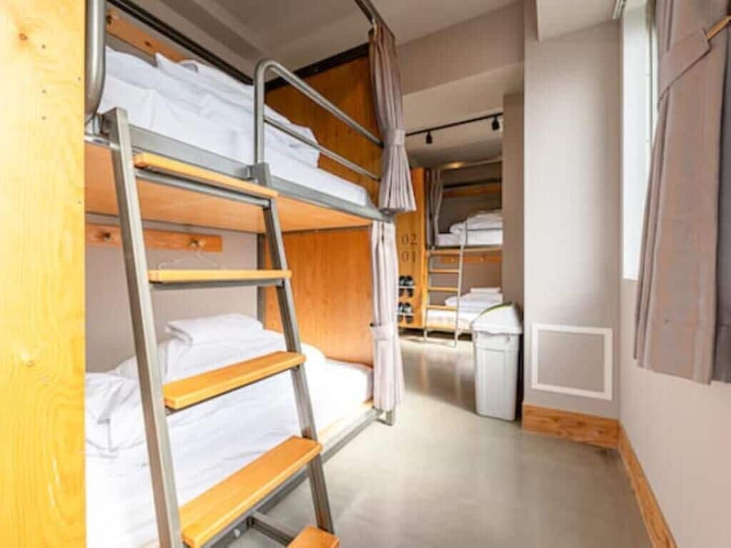 A dormitory room with bunk beds, a ladder, a small kitchenette, and a curtain.