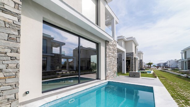 A modern poolside area with a stone wall and glass doors, a clear sky, and a view of neighboring houses.