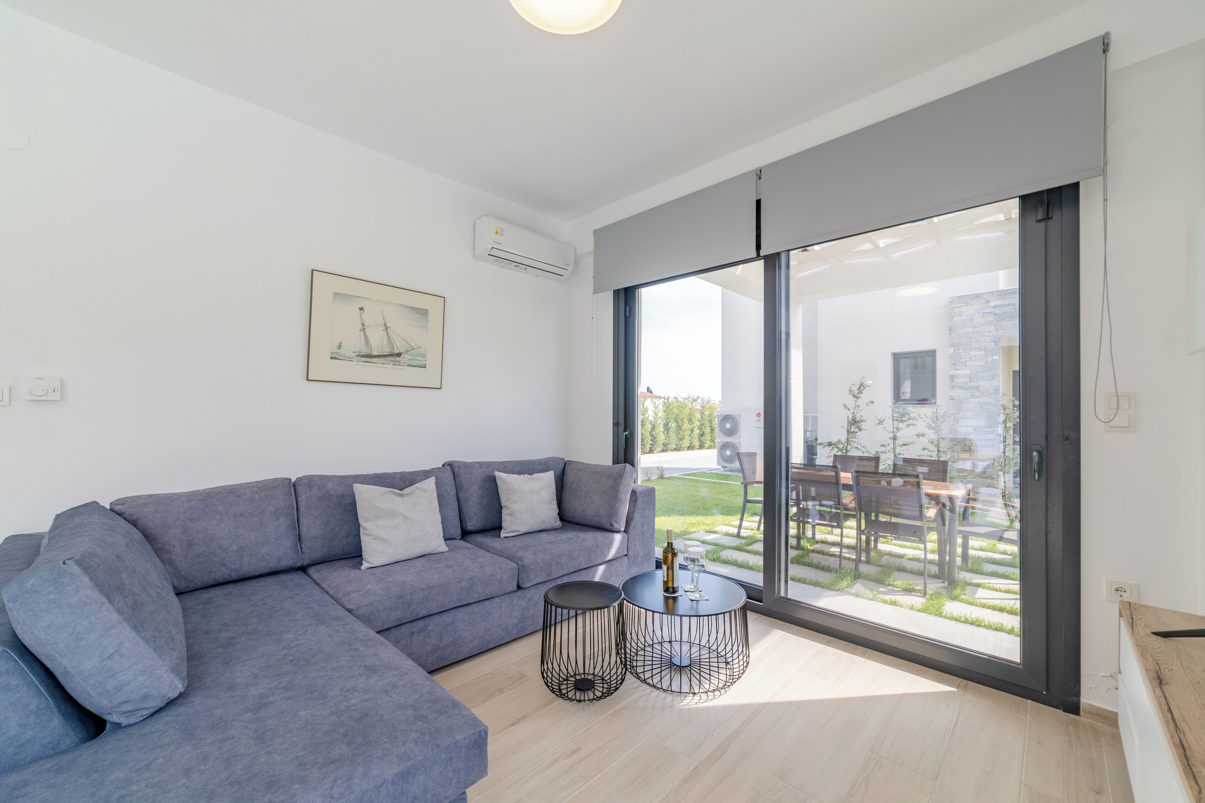 A modern living room with a grey sectional sofa, two black wireframe coffee tables, and a large sliding glass door leading to an outdoor patio with a dining set.