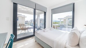A modern bedroom with a large bed, a balcony with a chair, and a view of buildings.