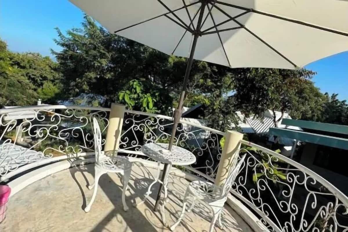 A balcony with white metal chairs and tables, an umbrella, and a view of trees and buildings.