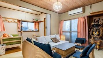A cozy room with a bed, a sofa, a chair, and a small table. There is a window with curtains, a hanging plant, and a wall-mounted shelf.