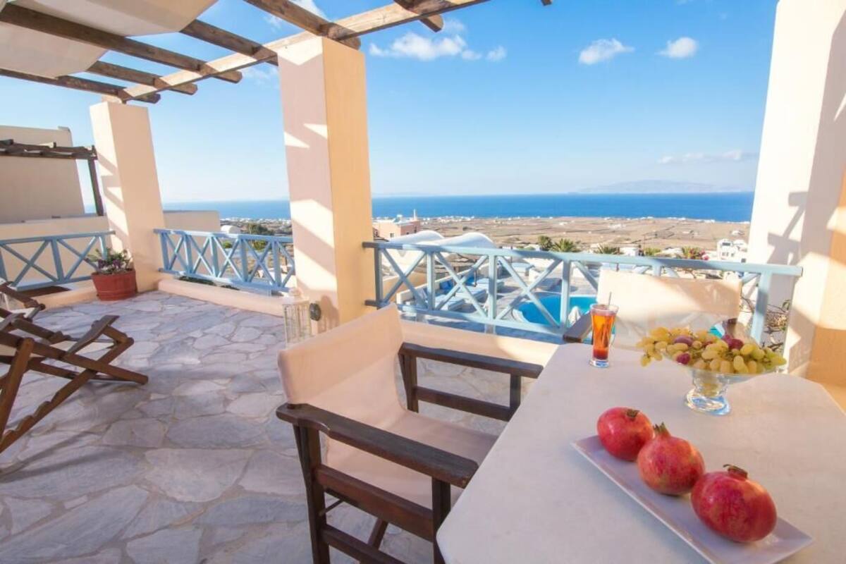Eine Terrasse mit Steinboden, Holzstühlen, einem Tisch mit Granatapfeln und einem Getränk, und Blick auf das Meer und die Ferne.