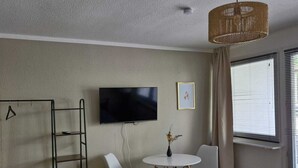 Un salon moderne doté d’un téléviseur à écran plat, d’une petite table ronde avec deux chaises, d’un luminaire suspendu en osier et d’une fenêtre avec des stores.