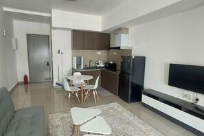 A modern living room with a sofa, a small dining table with chairs, a flat-screen TV mounted on the wall, and a kitchen area with cabinets and appliances. - Shahdila @ Alinea (Shah Alam)