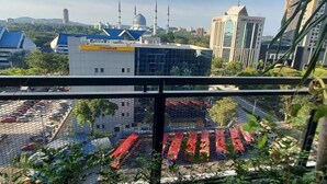 A view from a balcony with a cityscape in the background, including a mosque with minarets, a modern building with a sign, and a parking lot with red cars. - Shahdila @ Alinea (Shah Alam)
