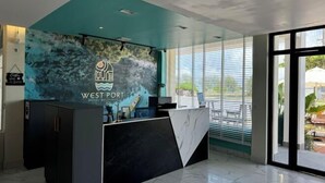 Un área de recepción de diseño moderno, con una pared verde azulado que muestra un logo, una barra de mármol y vistas amplias a través de grandes ventanales.