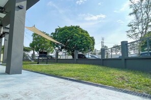 An outdoor area with a concrete floor, a grassy patch, a tree, and a metal fence. - Corner House Puchong 18 pax BBQ Kid Friendly (Puchong)