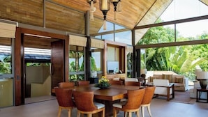 A spacious dining area with a wooden table and wicker chairs, large windows offering a view of lush greenery, and a modern interior design with a high ceiling. - Purana Residence (Koh Samui)