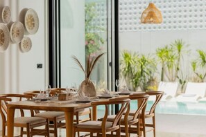 Un coin repas avec une table en bois dressée pour quatre personnes, entourée de chaises, et offrant une vue sur une piscine et la verdure environnante.
