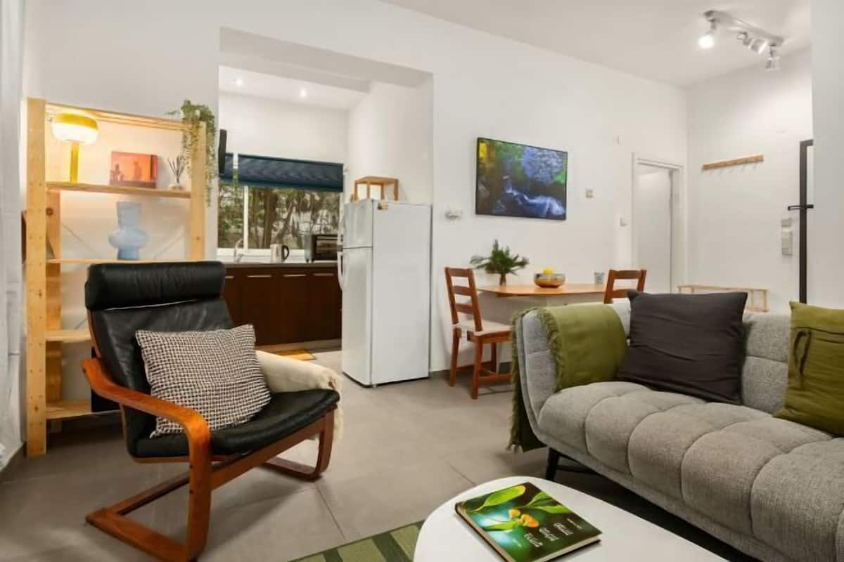 A modern living room with a sofa, a chair, a dining table with chairs, a bookshelf, and a television.