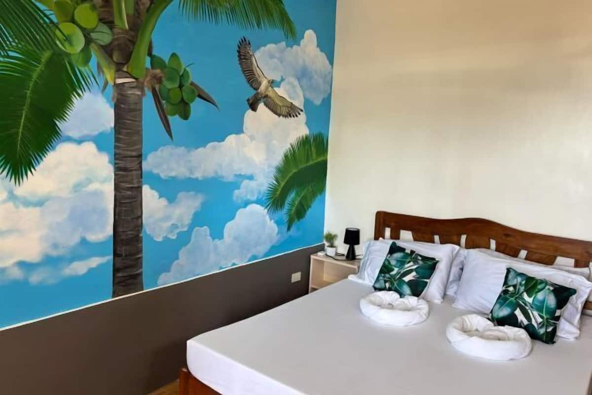 A bedroom with a bed, pillows, a nightstand, a lamp, and a wall mural of a palm tree and birds.