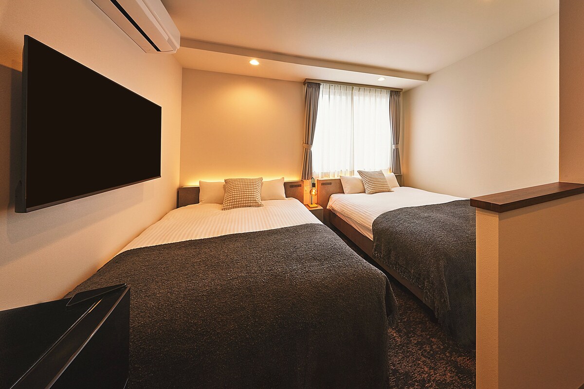 A hotel room with two beds, a flat-screen TV, and a window with curtains.