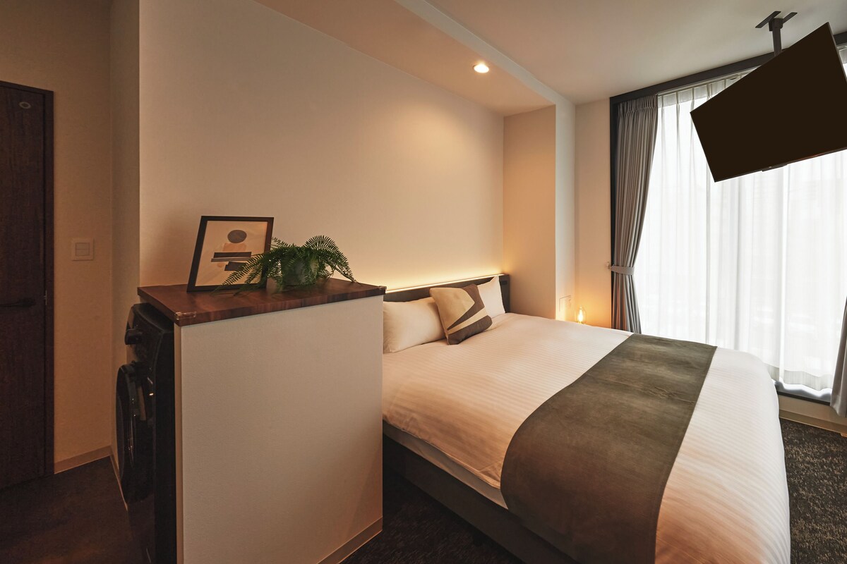 A hotel room with a bed, a desk with a lamp, a TV, and a window with curtains.