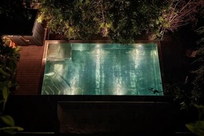 A rectangular swimming pool with a wooden deck surrounding it, surrounded by dense foliage.
