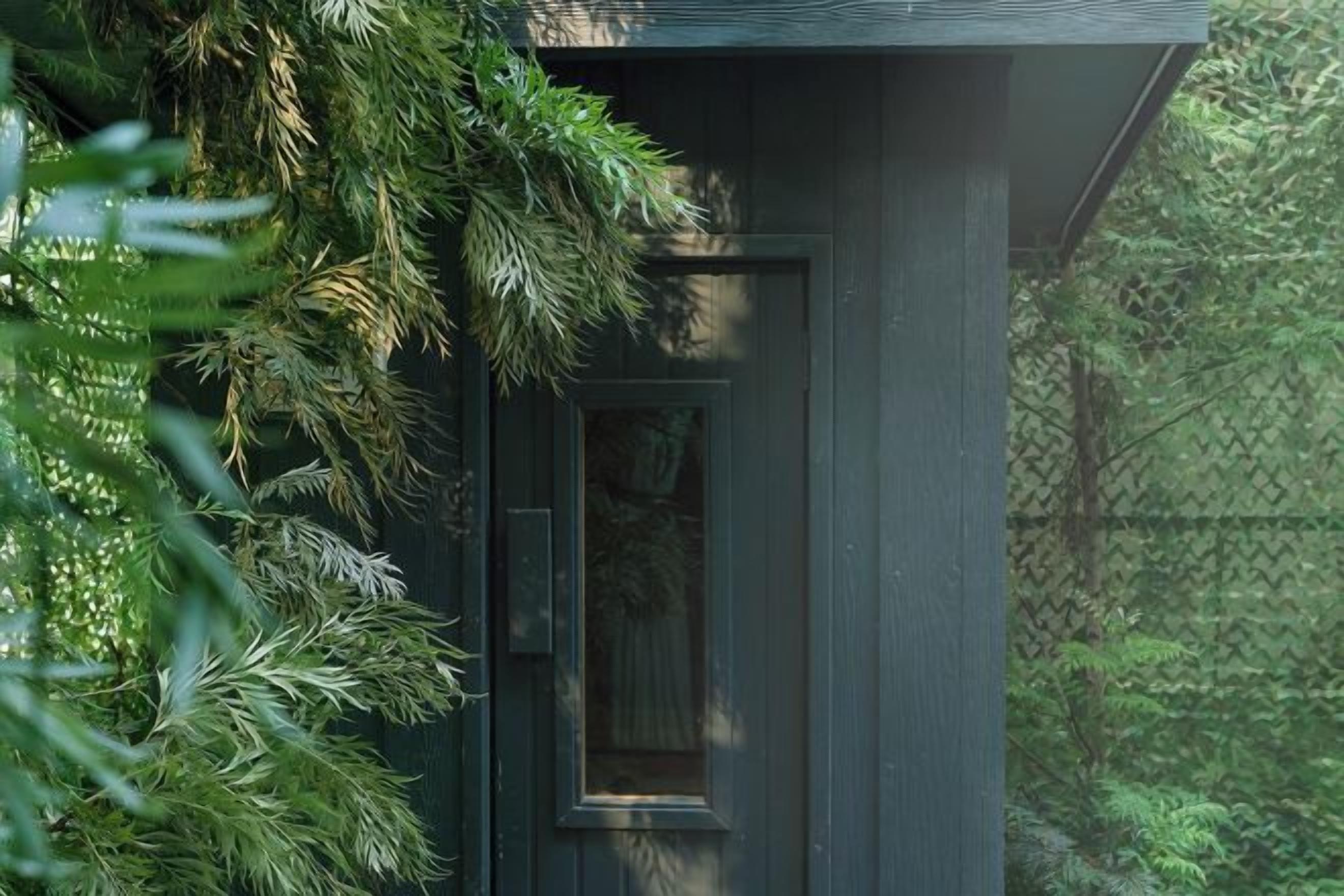 A dark-colored wooden structure with a door and window, surrounded by dense green foliage.