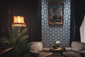 Una habitación con papel tapiz estampado, una obra de arte enmarcada, una mesa con un jarrón y dos sillones.