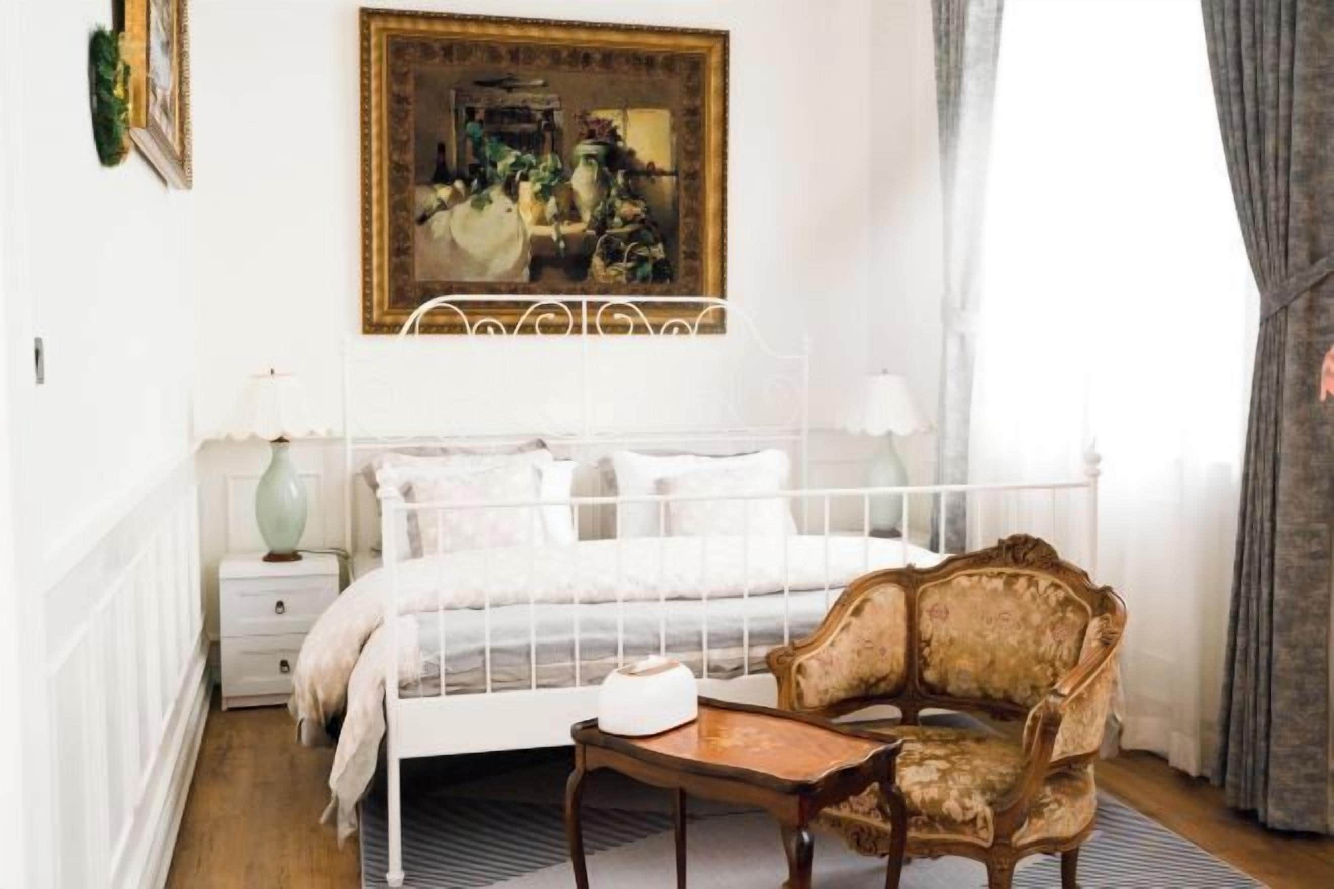 A bedroom with a white metal bed frame, a wooden chair, a bedside table, and a painting on the wall.