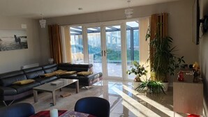 Een woonkamer met een bank, salontafel en eettafel. Er is een groot raam met gordijnen, een plant en uitzicht op het buiten.
