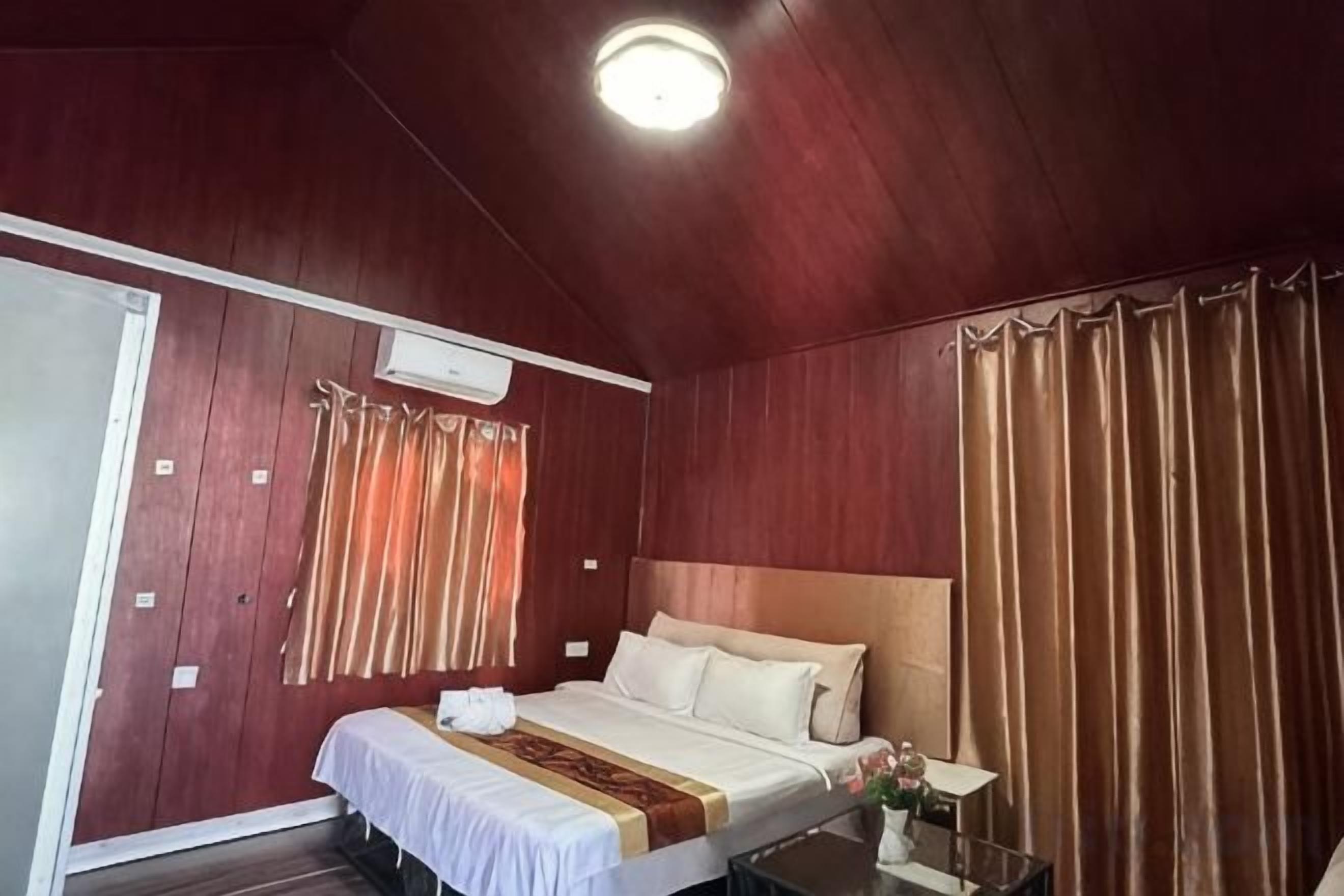 A hotel room with a wooden ceiling, a bed with white linens, a bedside table with a lamp, and a curtain on the window.
