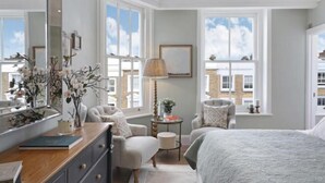 A bedroom with a large bed, two armchairs, a dresser, a mirror, and a window with a view of buildings. - 317 Grand Maisonette (London)